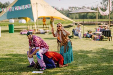 Saldus, Latvia - July 19, 2025 - People dressed in hippie-style clothing playfully posing and enjoying outdoor fun at a colorful summer festival with tents, decorations, and relaxed vibes.