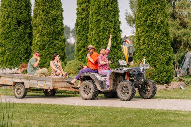 Saldus, Latvia - July 19, 2025 - A group of people in colorful hippie outfits enjoy a ride on a trailer pulled by an ATV during a fun summer festival surrounded by greenery and good vibes.