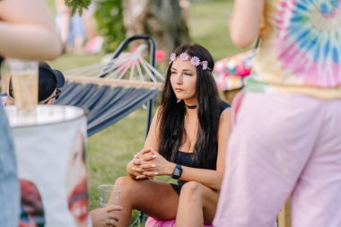 Saldus, Latvia - July 19, 2025 - A young woman in a flower crown sits pensively at a hippie festival, surrounded by people in colorful clothing, drinks, and relaxed summer vibes.