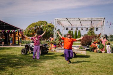 Saldus, Latvia - July 19, 2025 - Two men in colorful hippie clothes with microphones perform joyfully on a lawn stage at an outdoor festival decorated with ribbons, flowers, and bohemian props.