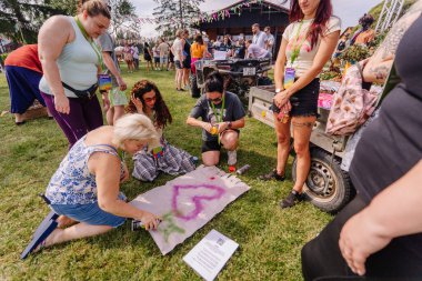 Saldus, Latvia - July 19, 2025 - People gather on grass at a hippie festival, creating spray-painted art on fabric in a colorful, creative workshop surrounded by music, laughter, and decorations.