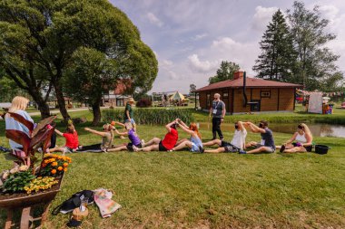 Saldus, Latvia - July 19, 2025 - A group of blindfolded people at a hippie festival sit in a line outdoors, passing water cups over their heads as part of a fun summer team-building challenge.
