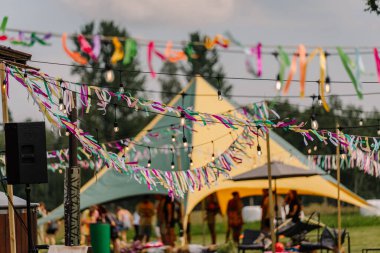 Saldus, Latvia - July 19, 2025 - Colorful streamers and hanging lights decorate an outdoor hippie festival with tents and blurred people in the background, creating a vibrant, festive atmosphere.