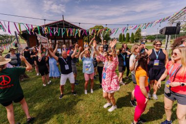 Saldus, Latvia - July 19, 2025 - A joyful group of people dance and celebrate together at an outdoor hippie-style festival, surrounded by colorful ribbons and festive decorations.