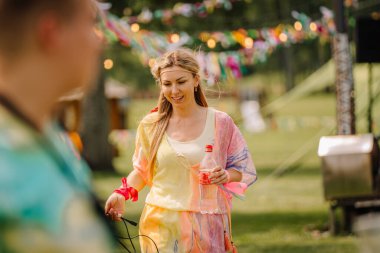 Saldus, Latvia - July 19, 2025 - A smiling woman in colorful hippie-style clothing holds a bottle and cable, enjoying a sunny outdoor festival with vibrant decorations in the background.