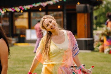 Saldus, Latvia - July 19, 2025 - A cheerful woman in a colorful boho outfit smiles at the camera during an outdoor summer hippie festival, holding a bottle, surrounded by vibrant decorations.