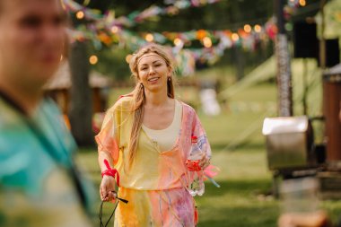 Saldus, Latvia - July 19, 2025 - A smiling woman in colorful hippie-style clothing holds a bottle and cable, enjoying a sunny outdoor festival with vibrant decorations in the background.