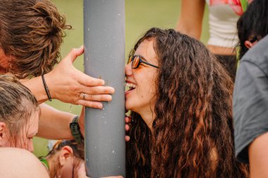 Saldus, Latvia - July 19, 2025 - Close-up of smiling young woman with wet hair and sunglasses taking part in a fun, water-based team challenge during an outdoor hippie festival.