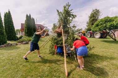Saldus, Latvia - July 19, 2025 - People build a makeshift shelter from branches during a fun outdoor game at a hippie festival, while one man playfully pours water over the structure.