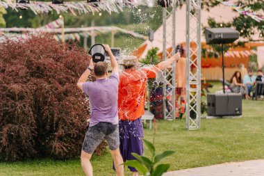 Saldus, Latvia - July 19, 2025 - Man splashes water on another man holding a microphone during a playful moment at a hippie festival. Laughter, spontaneity, and summer fun fill the air.