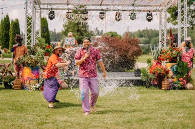 Saldus, Latvia - July 19, 2025 - A performer with a microphone gets splashed with water on stage during a lively hippie festival surrounded by flowers, decorations, and festive energy.