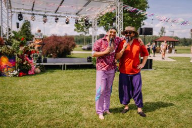 Saldus, Latvia - July 19, 2025 - Two cheerful men in colorful outfits hold microphones on an outdoor stage at a hippie festival, smiling and enjoying the lively summer event.