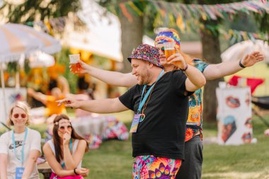 Saldus, Latvia - July 19, 2025 - Two cheerful men in colorful hippie outfits dance with drinks in hand at an outdoor festival, while smiling people watch and enjoy the laid-back atmosphere.