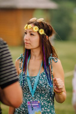 Saldus, Latvia - July 19, 2025 - Young woman at a hippie festival wearing a flower crown, sunglasses, braided hair with colorful extensions, floral dress, and event badge, enjoying the outdoors.