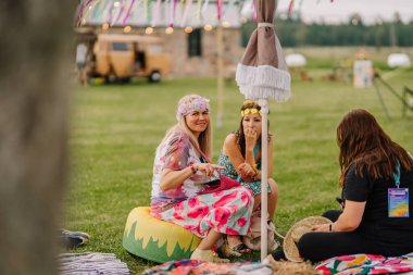 Saldus, Latvia - July 19, 2025 - Three women dressed in colorful boho outfits sit on grass under a sun umbrella at a hippie festival, chatting and relaxing in a joyful, peaceful outdoor atmosphere.