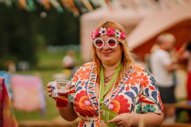 Saldus, Latvia - July 19, 2025 - Smiling woman wearing colorful floral dress and flower sunglasses holds a drink at a vibrant outdoor hippie festival, surrounded by decorations and people.