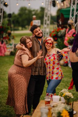 Saldus, Latvia - July 19, 2025 - Three cheerful friends pose together at a colorful outdoor hippie festival, dressed in retro outfits and smiling in a vibrant summer atmosphere.