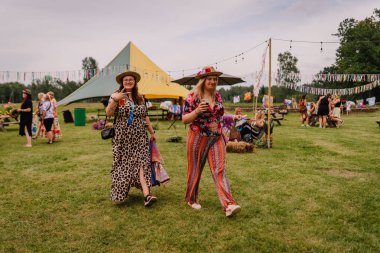 Saldus, Latvia - July 19, 2025 - Two smiling women in colorful hippie-style outfits walk across a vibrant outdoor festival grounds with tents, decorations, and people enjoying the summer vibe.