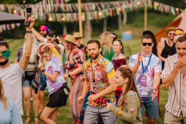 Saldus, Latvia - July 19, 2025 - A vibrant group of people dance enthusiastically at a colorful outdoor hippie festival, dressed in retro and tie-dye outfits, enjoying music and freedom.