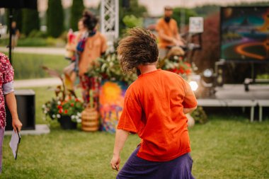 Saldus, Latvia - July 19, 2025 - A man in an orange shirt dances freely with his hair swinging at an outdoor hippie festival, near a colorful stage and psychedelic decorations.
