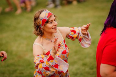 Saldus, Latvia - July 19, 2025 - Smiling woman in a colorful retro outfit and headband dancing joyfully on the grass during an outdoor hippie festival.