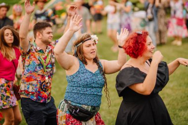 Saldus, Latvia - July 19, 2025 - Joyful group of people dancing outdoors at a hippie festival. Bright outfits, raised hands, smiles, and carefree vibes highlight the celebration atmosphere.