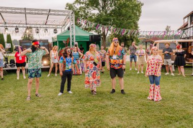 Saldus, Latvia - July 19, 2025 - A group of people dressed in colorful, retro hippie outfits stand on grass at an outdoor festival, surrounded by decorations, music, and cheering spectators.