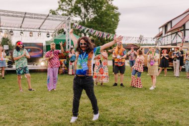 Saldus, Latvia - July 19, 2025 - A man in a hippie costume raises his arms while others cheer and clap during a colorful outdoor hippie-themed festival with music, fun, and vintage vibes.