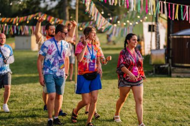 Saldus, Latvia - July 19, 2025 - Group of people walking and chatting at a hippie festival, dressed in colorful tie-dye shirts and boho outfits, surrounded by festive ribbons and warm sunlight.