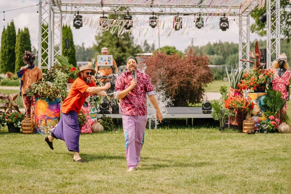 Saldus, Latvia - July 19, 2025 - Man splashes water on performer with a microphone during a fun moment on stage at a hippie festival, surrounded by flowers, decorations, and laughter.