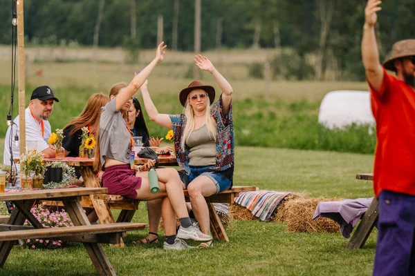 Saldus, Latvia - July 19, 2025 - People enjoy a relaxing moment at a hippie festival, sitting at wooden picnic tables with sunflowers, raising their hands and soaking in the laid-back vibe.