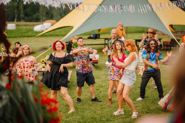 Saldus, Latvia - July 19, 2025 - A joyful group of people dances at an outdoor hippie festival, dressed in colorful boho outfits, under festive ribbons and tents on a sunny day.