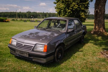 Saldus, Latvia - July 19, 2025 - An old, weathered Opel sedan parked on green grass under a tree in a rural setting. The vintage car shows signs of age, rust, and wear.