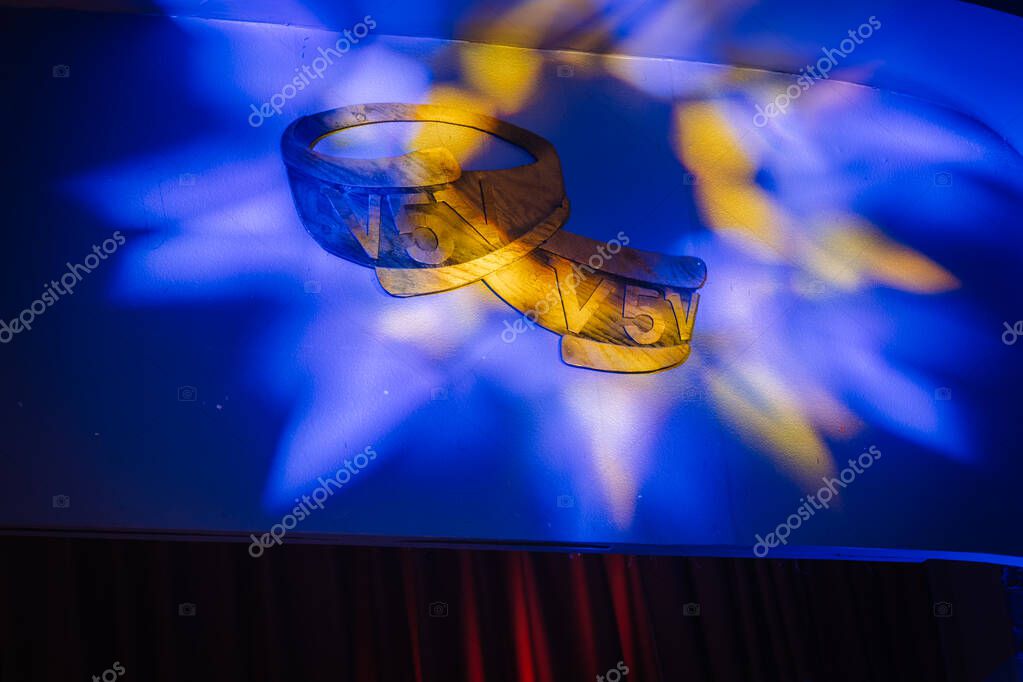 Valmiera, Latvia - March 7, 2025 - Decorative wooden V5V emblem mounted on a wall, illuminated with colorful blue and yellow stage lighting, likely part of an event or ceremony decor