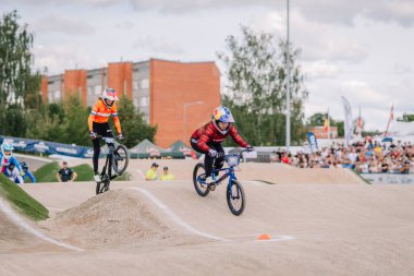 Valmiera, Letonya - 12 Temmuz 2025: BMX Avrupa Şampiyonası. Kırmızı ceketli erkek BMX sürücüsü, toprak pistte kalabalıkla yarışıyor.