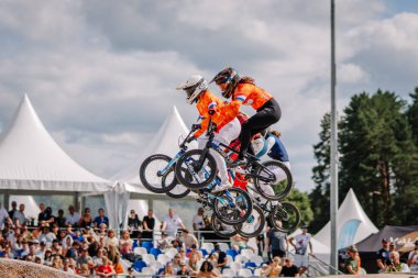 Valmiera, Letonya - 12 Temmuz 2025: BMX Avrupa Şampiyonası. Turuncu formalı kadın BMX sürücüleri kalabalıkla bisiklet yarışında havada süzülüyorlar.