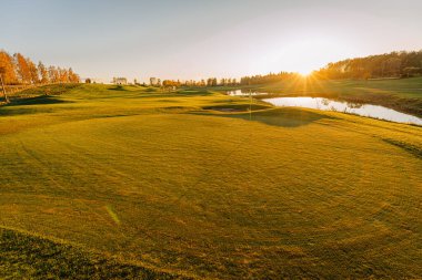 Sonbahar boyunca altın gün batımında bir golf sahasının manzarası, sonbahar renklerinde ağaçlar ve sakin bir su yansıması. Huzurlu ve huzurlu manzara.