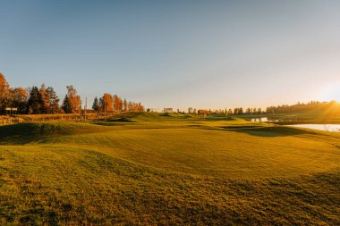 Sonbahar boyunca altın gün batımında bir golf sahasının manzarası, sonbahar renklerinde ağaçlar ve sakin bir su yansıması. Huzurlu ve huzurlu manzara.