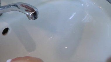 Ellerin bir musluğun altına yerleştirilmesi, yıkama işlemini yakalamak için yavaşça yakınlaşması, su akışını ve el hareketlerini vurgulaması, sahne boyunca temiz ve parlak bir ortam yaratması.