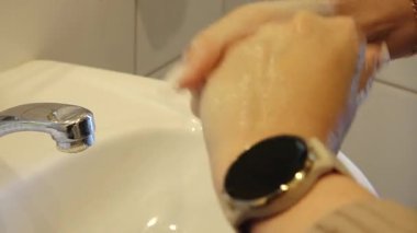 Eller aktif olarak akan suyun altında yıkanır, sabun köpük oluşturur, hijyen ve temizliğin önemini vurgular, kamera detaylı yıkama işlemini yakalamak için yakınlaşır