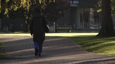 Valmiera, Letonya - 17 Ekim 2025: Siyah paltolu bir kadın parktaki eğri bir yolda yürüyor, kamera onu takip ediyor.