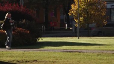 Valmiera, Letonya - 17 Ekim 2025: Kadın figürü parkta küçük bir köpekle yürüyor, kamera onu renkli sonbahar yaprakları ve güneş ışığı boyunca takip ediyor