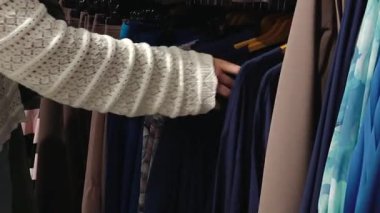 Valmiera, Letonya - Ekim 06, 2025: Beyaz kazaklı bayan alışverişçi elbise raflarını, kamera tavalarını geziyor ve seçtiği kıyafetleri canlı renklerle gösteriyor