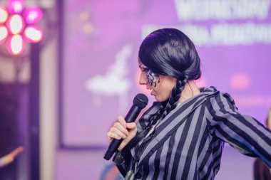 Koyu renk saçlı ve benzersiz makyajlı bir kadın, elinde mikrofonla sahnede coşkulu bir şekilde performans sergiliyor. Renkli ışıklar ve coşkulu bir ortam yaratıyor.