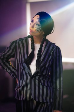 Çizgili takım elbiseli, özenli yüz boyalı, kendine güvenen bir kadın model çağdaş bir ortamda poz veriyor, eşsiz stilini ve sanatsal yeteneğini vurguluyor.