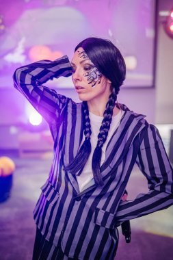 Örgülü saçlı, şık çizgili takım elbiseli, renkli ışıklarla canlı bir ortamda poz veren, sanatsal makyajı ve benzersiz moda tercihlerini vurgulayan bir kadın model.