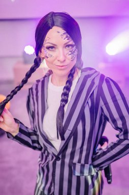 Çizgili takım elbiseli ve detaylı yüz boyalı bir kadın renkli ışıklarla çevrili bir etkinlikte güven içinde duruyor. Yaratıcılık ve bireyselliği dinamik bir ortamda vurguluyor.