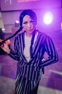 Kendine güvenen, örgülü saçlı, çizgili takım elbiseli ve eşsiz makyajlı bir kadın sanatçı renkli sahne ışıkları altında canlı bir atmosfer yaratıyor.