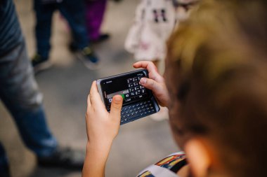 Riga, Letonya - 24 Ekim 2025: Genç çocuk, insanlarla dolu canlı bir açık hava ortamında fiziksel klavye içeren akıllı telefonla etkileşime girdi