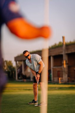 Erkek golfçü, arka planda kırsal bir kulüp evi bulunan canlı yeşil bir sahaya odaklanarak rahat bir atmosfer ve sportif bir adanmışlık sergiliyor.